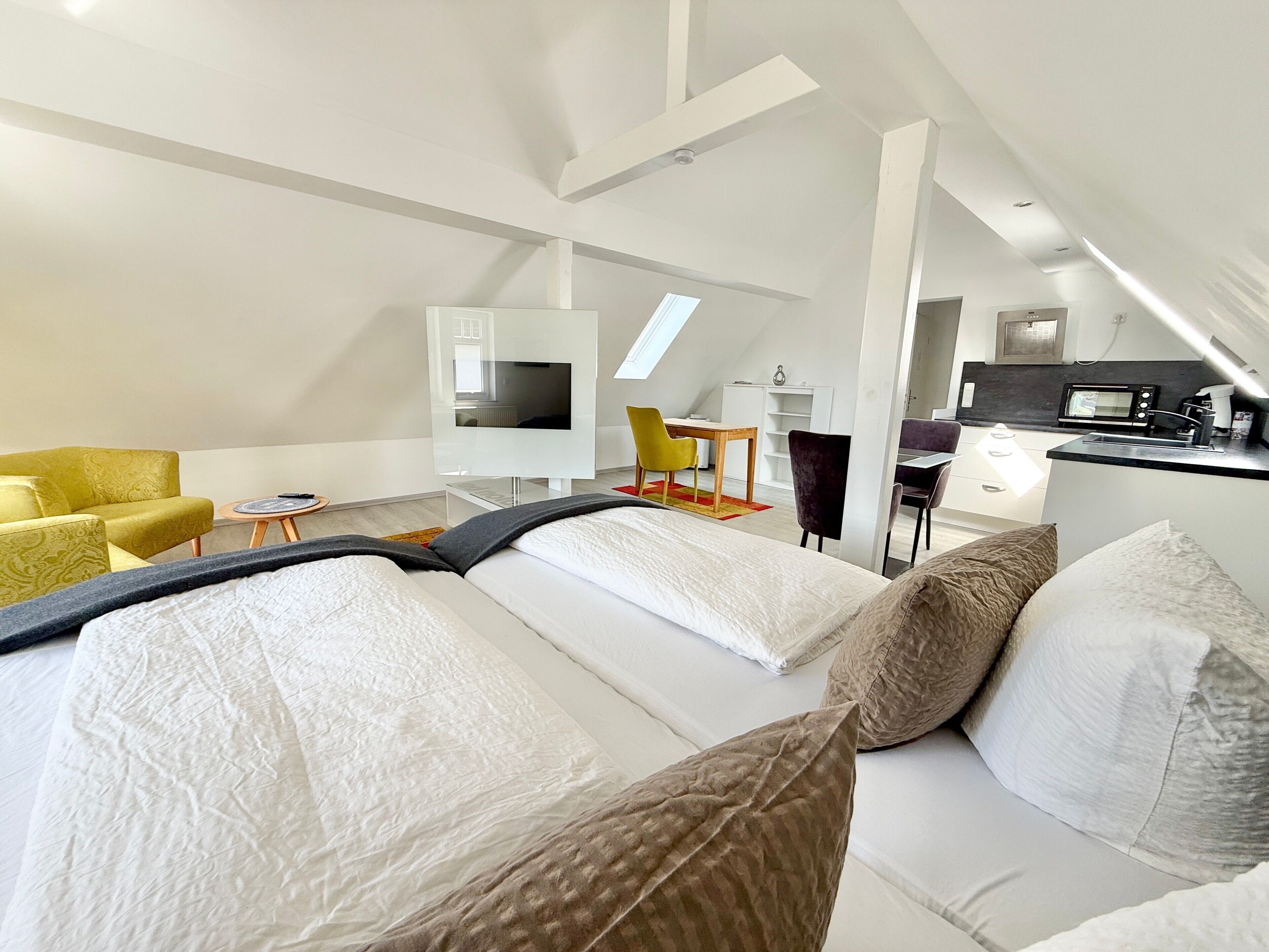 A modern hotel room with a loft-style ceiling, a bed with white linens, a yellow armchair, a desk with a chair, and a kitchenette area.