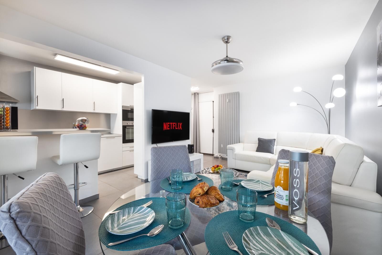Un salon moderne doté d’un téléviseur à écran plat, d’un coin repas avec une table en verre et d’une cuisine avec des meubles blancs.