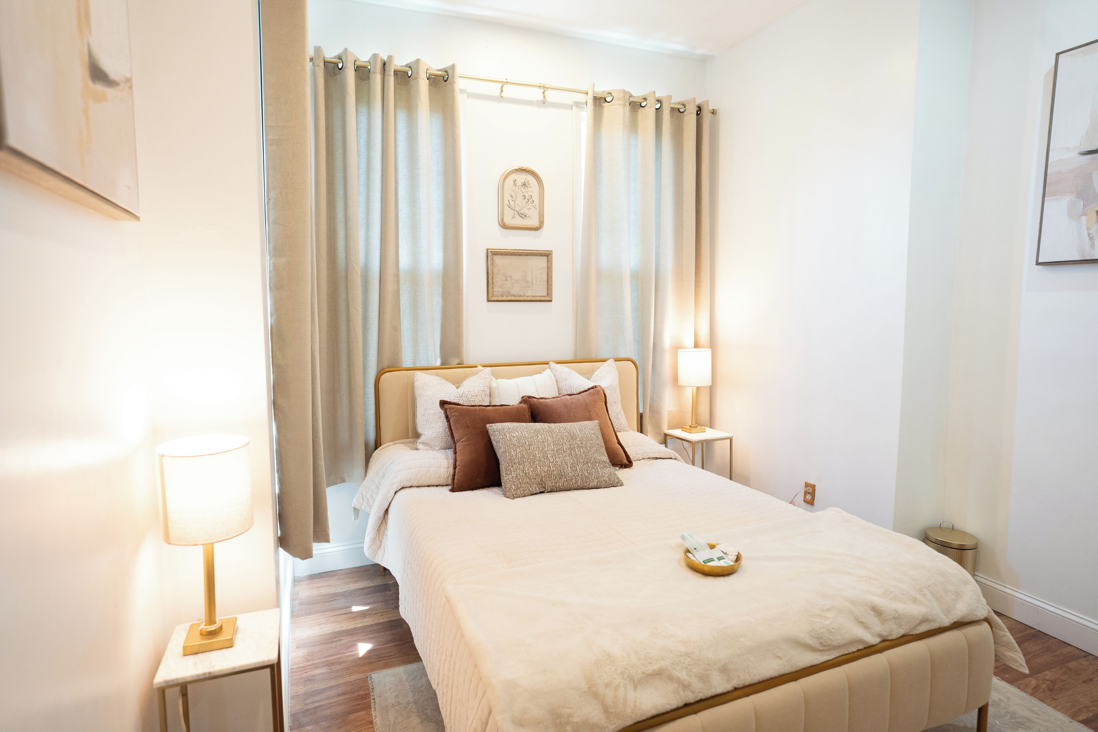 Un dormitorio con cama, cortinas, lámparas de noche, mesita de noche y un cuadro en la pared.