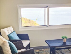A room with a view of the ocean through a window, a wicker chair with cushions, and a small table with a plant. - Betty's House, Ocean and Hillside View (Fajã da Ovelha)