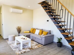 A modern living room with a grey sofa, wooden staircase, and a white air conditioner. - Betty's House, Ocean and Hillside View (Fajã da Ovelha)