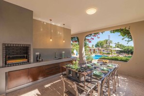 Eine moderne Küche mit eingebautem Grill, ein Essbereich mit Blick auf einen Swimmingpool und eine Terrasse mit Sitzgelegenheit.