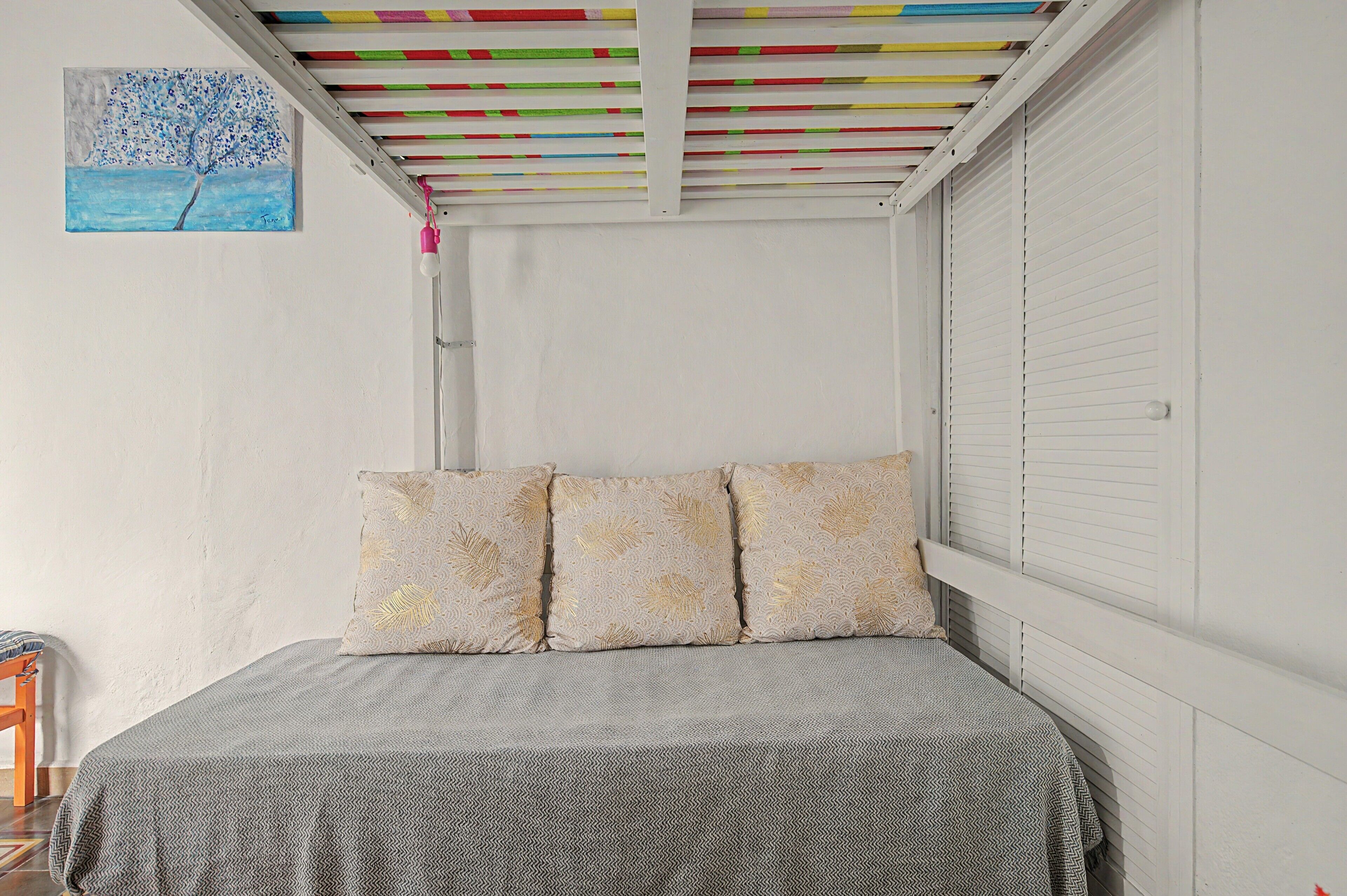 A bedroom with a bed, pillows, a painting, and a ceiling with colorful beams.