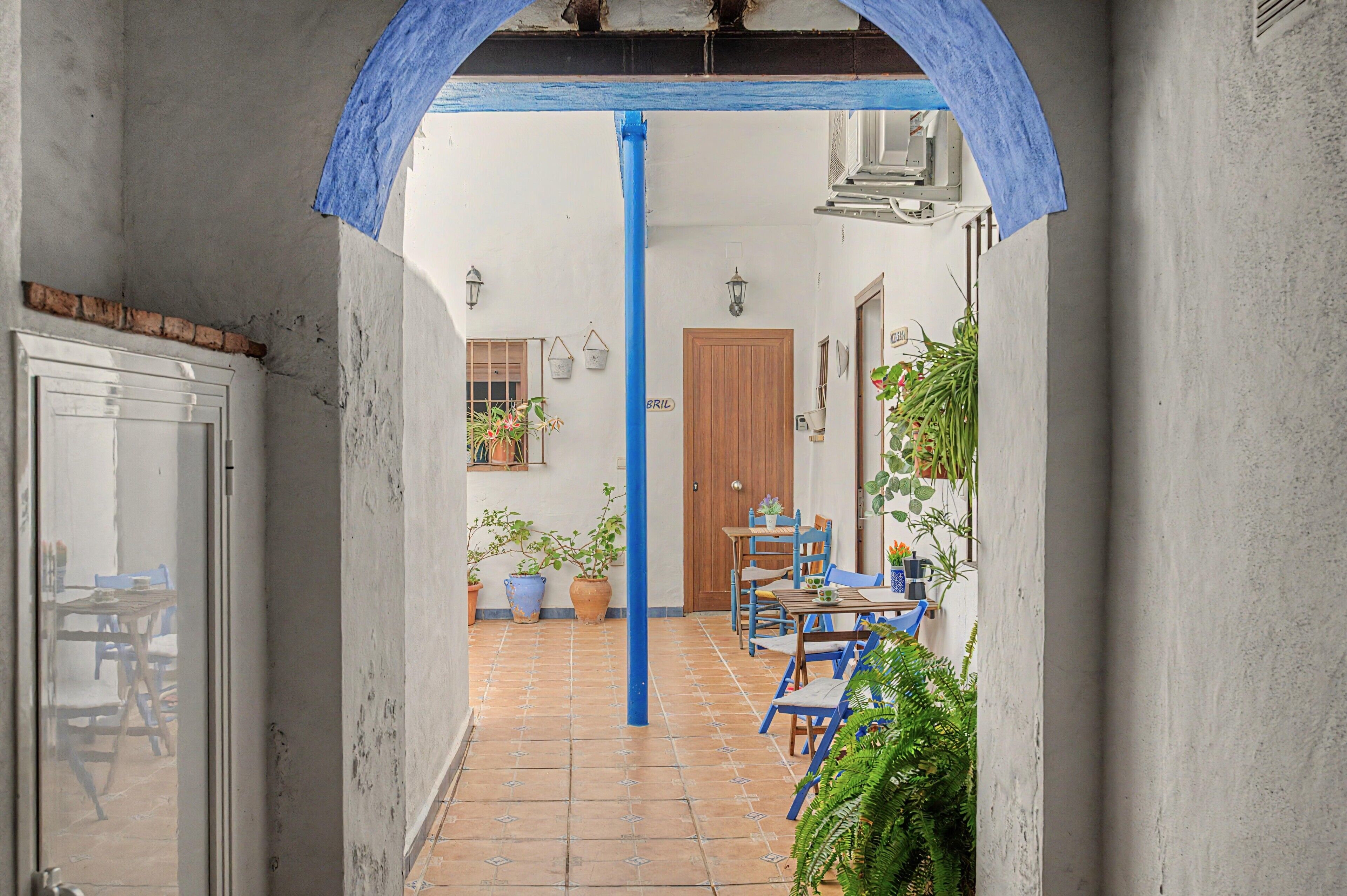 A narrow corridor with a tiled floor leading to a room with a wooden door, a table with chairs, and potted plants.