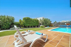 Un espace aménagé au bord de la piscine, avec un fauteuil de relaxation blanc, une serviette bleue et une piscine dont le fond est bordé d’un mur en pierre.