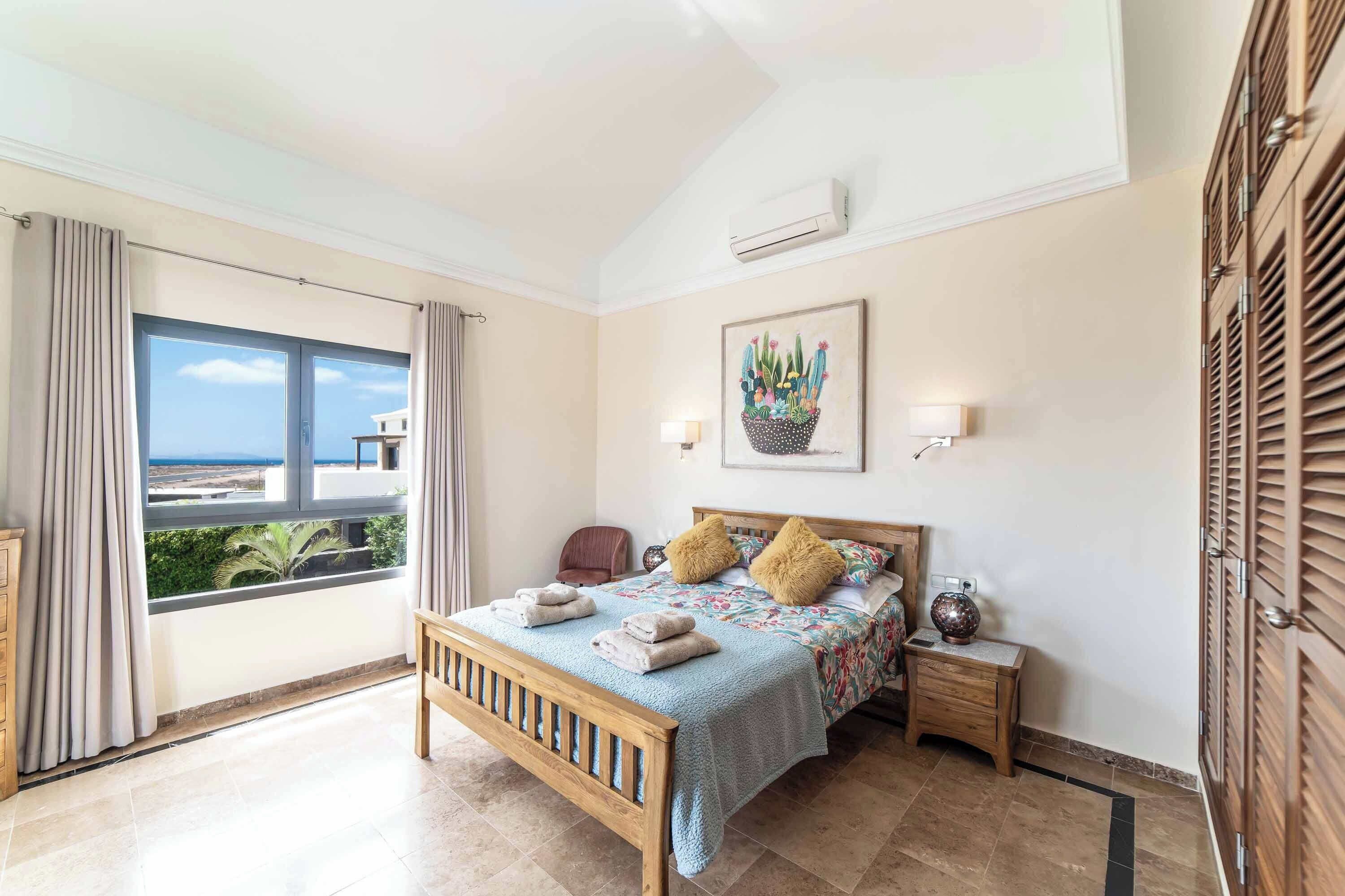 A bedroom with a bed, a nightstand, a lamp, a window with curtains, and a wall-mounted air conditioner.