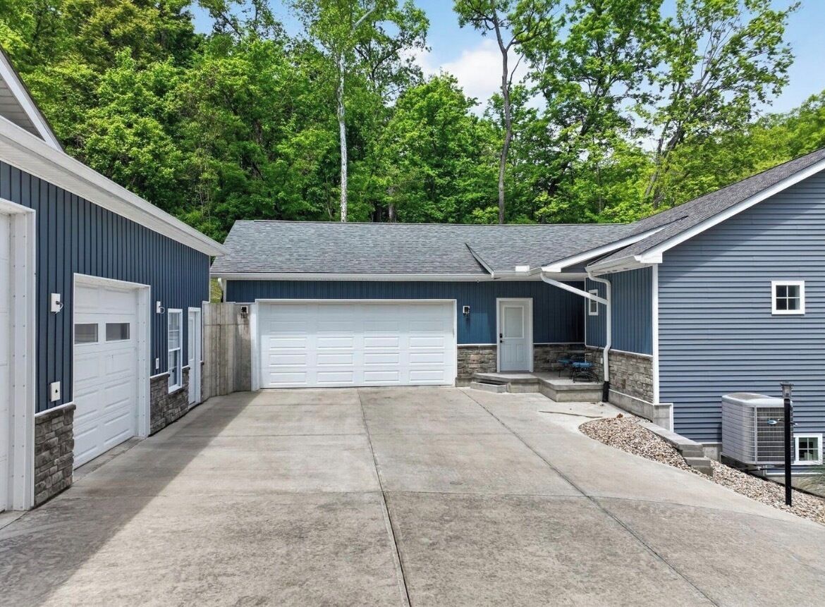 Un garage attenant à une maison, avec une allée en béton, un mur en pierre et une maison bleue dotée d’une porte de garage blanche.