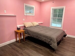 A bedroom with a bed, two windows with blinds, a bedside table, and a power outlet on the wall.