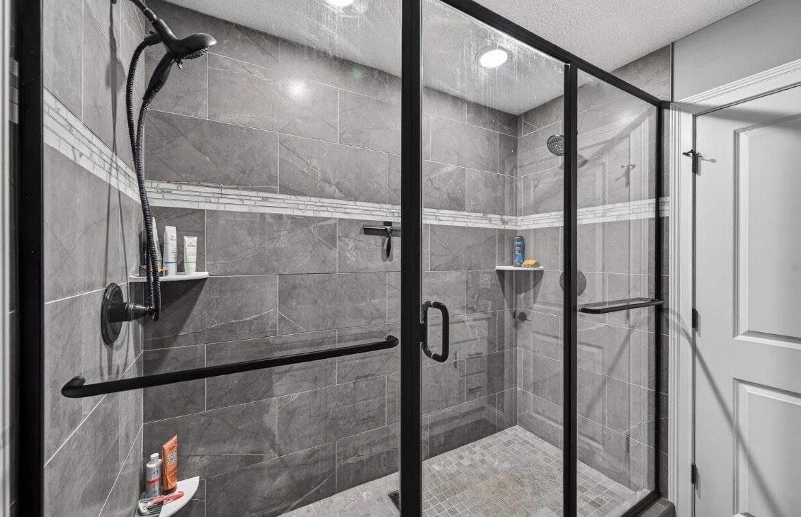 A glass-enclosed shower stall with a handheld showerhead, a towel rack, and a shelf with toiletries.