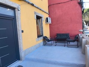 A building with a yellow exterior, a red wall, a black door, and a small window with an air conditioning unit. There are two black chairs and a table outside. - Casa Stone – Local Accommodation (Guarda)
