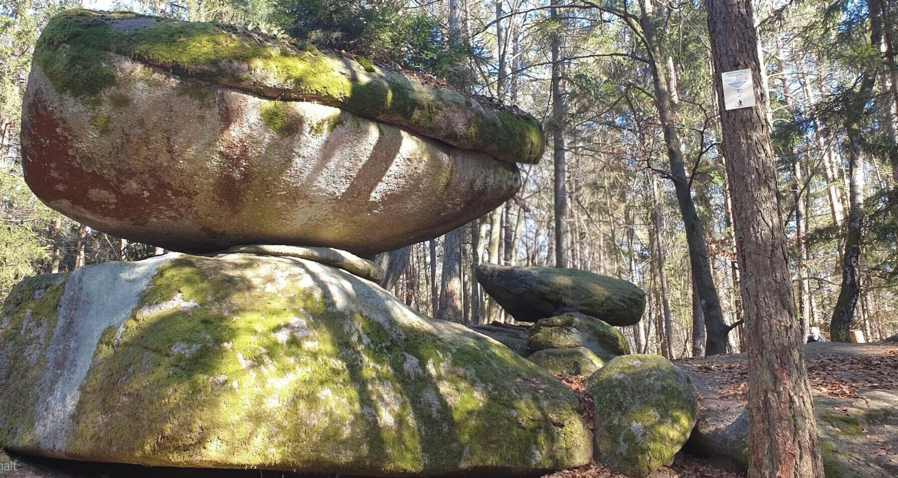 Un cadre naturel en plein air, avec de gros rochers recouverts de mousse et un panneau indiquant le début d’un sentier pédestre.