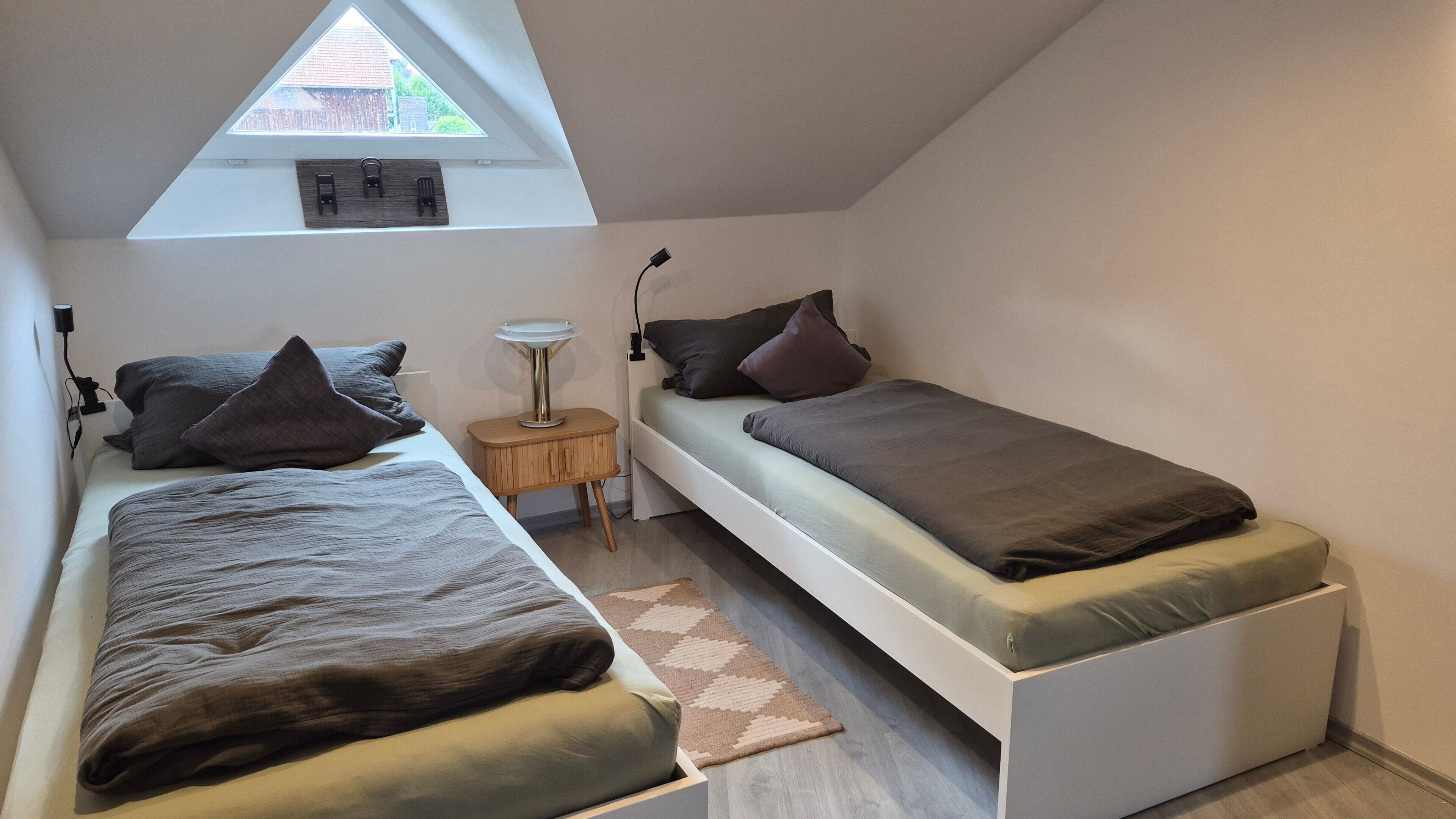 A small, modern bedroom with two single beds, a skylight, a bedside table, and a floor lamp.
