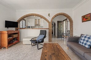 A living room with a television, a sofa, a coffee table, and a chair. There is a kitchen area with cabinets and a microwave. The room has a tiled floor and brick arches.