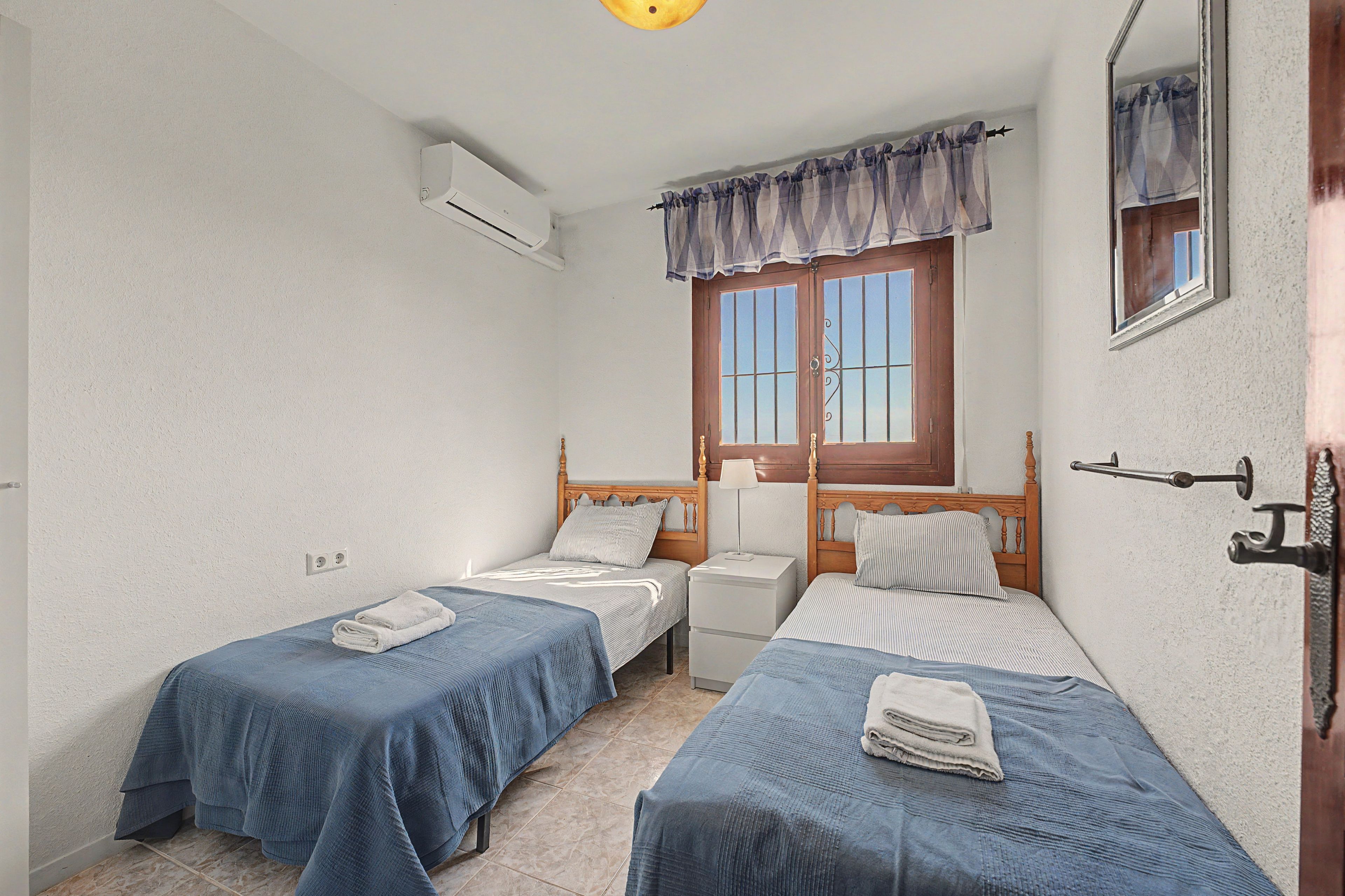 Une petite chambre propre, avec deux lits simples, une fenêtre avec des rideaux, une table de chevet et un appareil de climatisation.