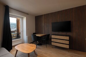 A modern living room with a wooden floor, a round table, a sofa, a TV mounted on a wall, and a window with a view of a cityscape. - K2 - Luxury Apartment: Terrace and View of the Slopes (Gérardmer)