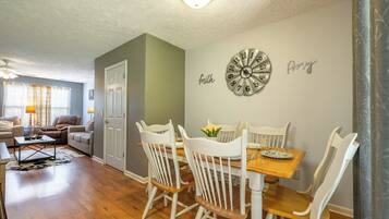 A dining area with a wooden table and white chairs, a wall clock, and a door leading to another room.