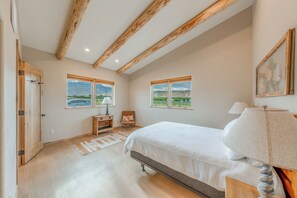 A bedroom with a bed, two windows with views, a wooden door, and a nightstand. - Peach Valley Vista - Sunset Views, Orchards & Wine Country Escape (Palisade)