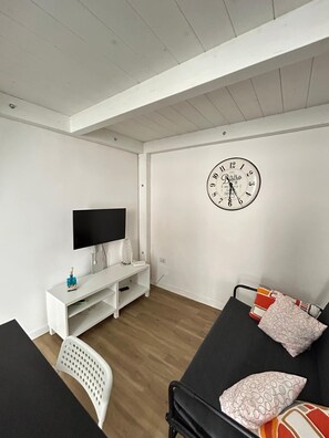 Ein modernes Wohnzimmer mit einem Flachbildfernseher, einer weißen Unterhaltungsanlage, einem schwarzen Sofa mit gemusterten Kissen und einer großen Wanduhr.