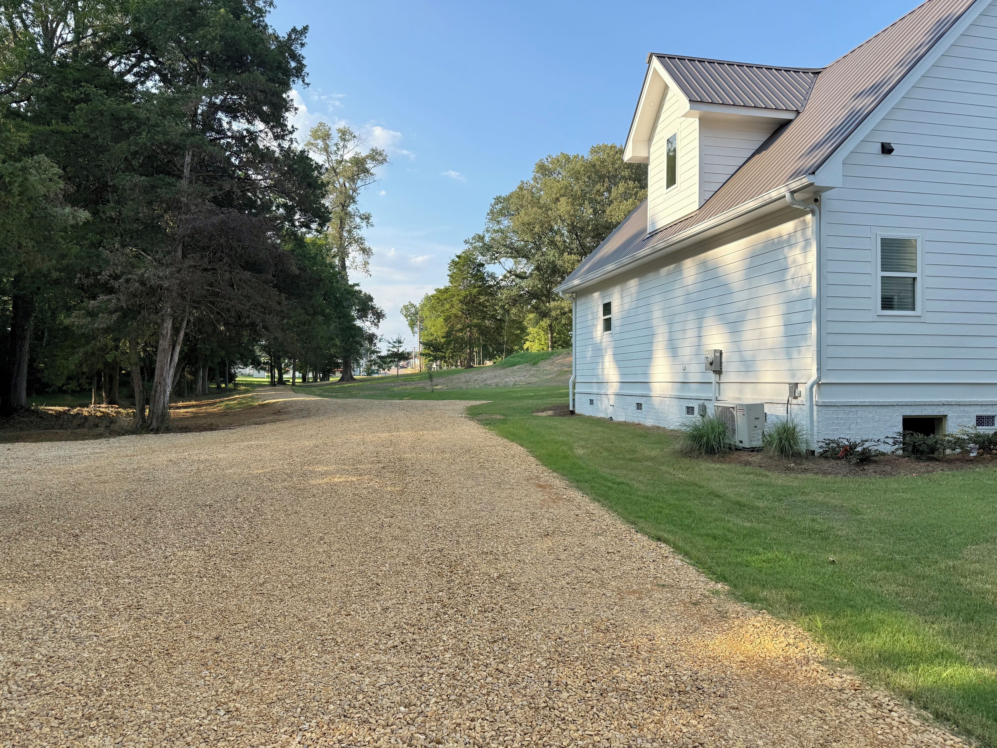 A gravel driveway leading to a white house with a sloped roof, surrounded by trees and a grassy area.