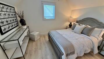 A bedroom with a bed, bedside tables, a nightstand, a window with blinds, and a wall-mounted artwork.