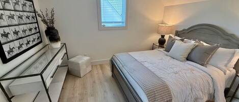 A bedroom with a bed, bedside tables, a nightstand, a window with blinds, and a wall-mounted artwork.