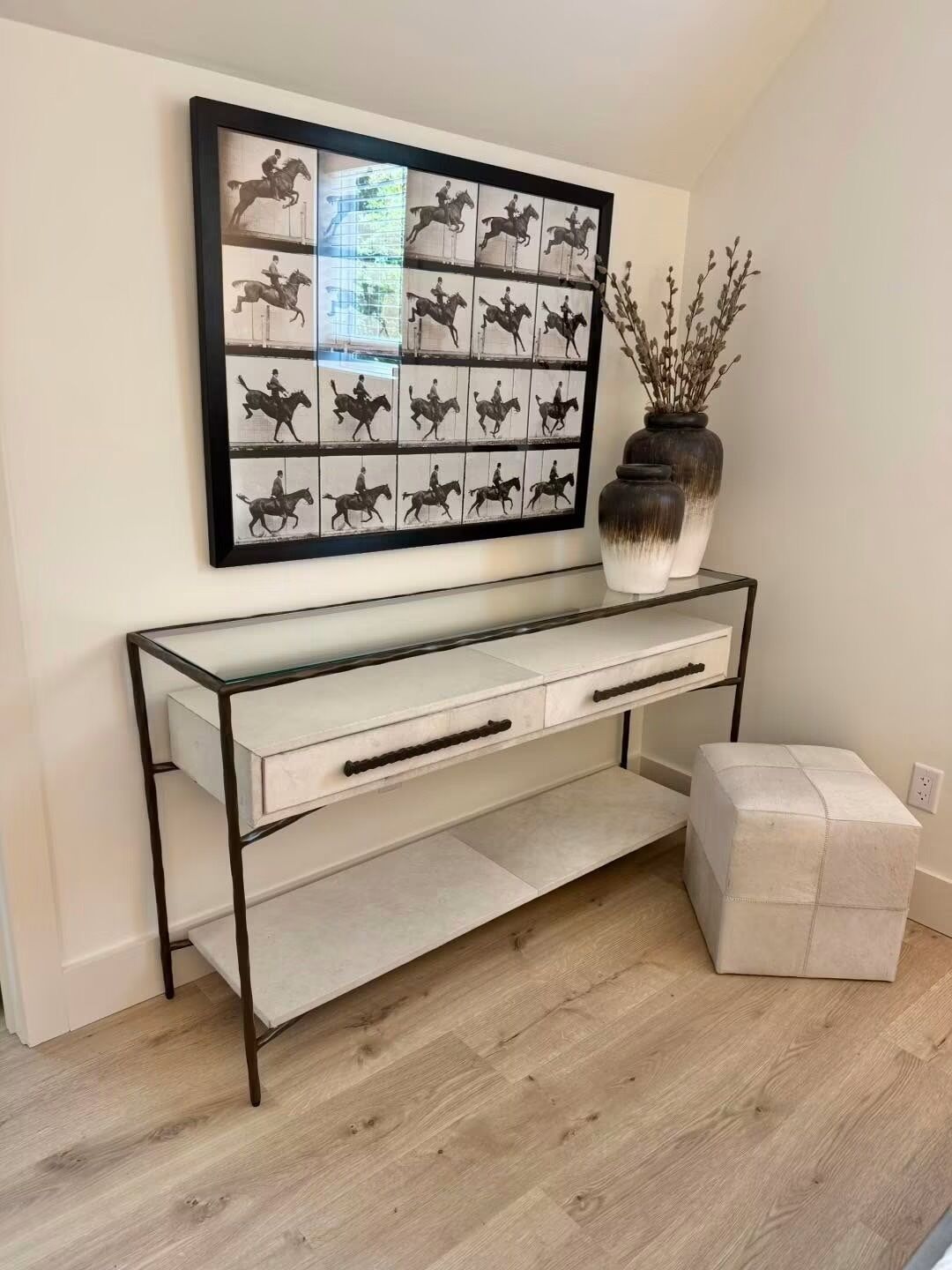 A modern console table with a glass top and metal frame, accompanied by a vase of dried branches, set against a plain wall.