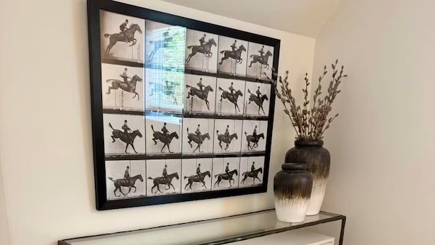 Een moderne console tafel met een glazen blad en metalen frame, met een vaas met gedroogde takken, voor een effen muur.