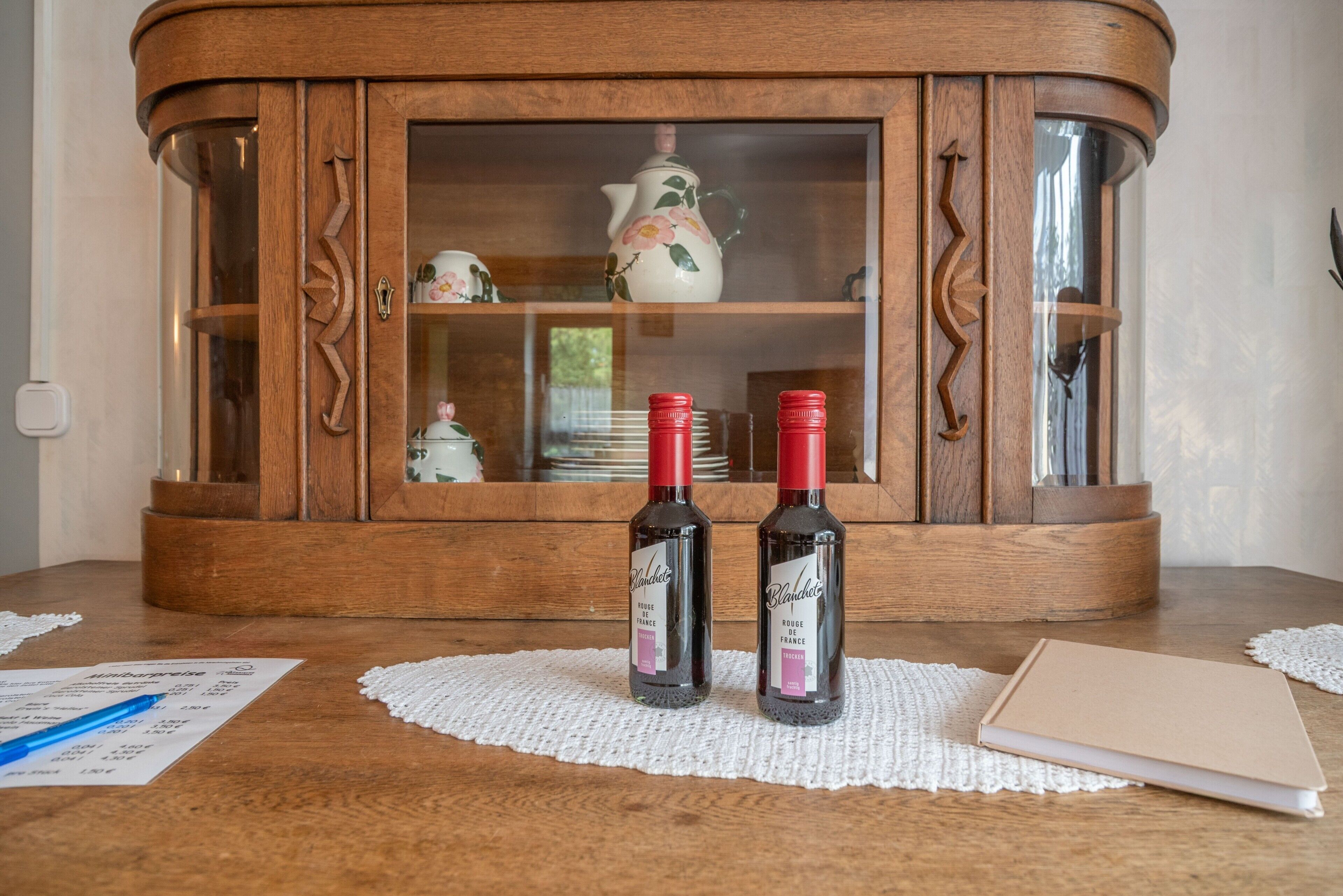 A wooden cabinet with glass doors, two bottles of wine, a receipt, and a notebook on a wooden table.