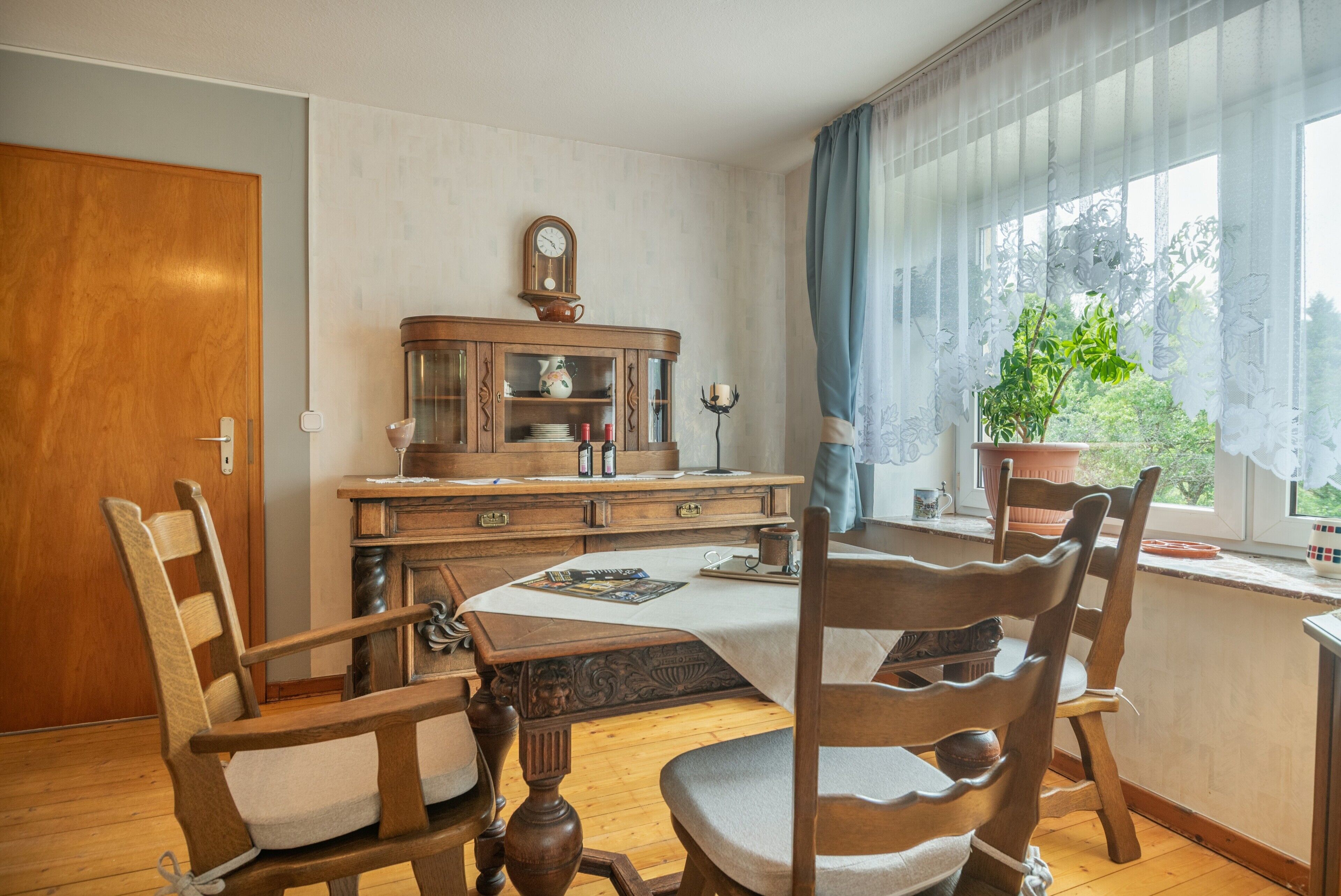 Ein traditioneller Speisesaal mit Holzmöbeln, einer Vitrine mit Glastüren und einem Fenster mit Vorhängen.