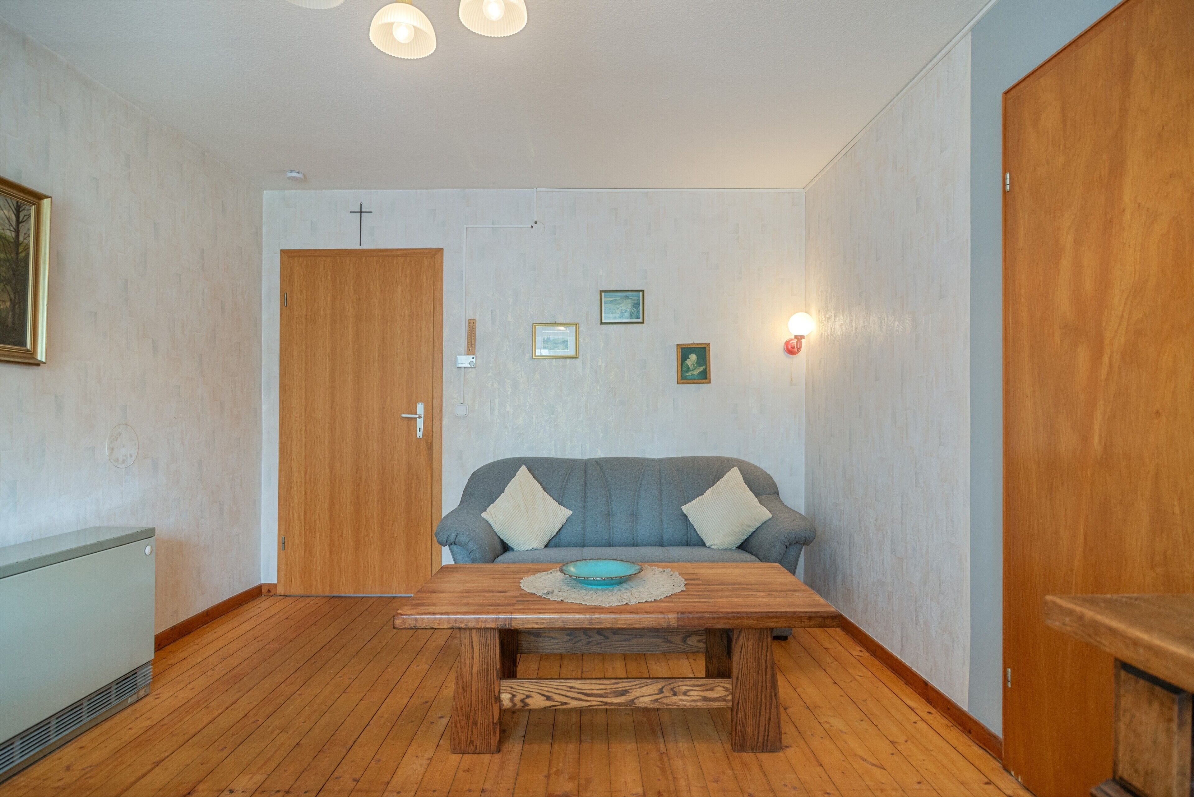 Ein Zimmer mit Holzboden, einem grauen Sofa mit Kissen, einem Holztisch und einer Tür.