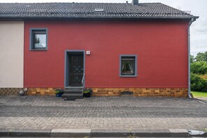 A red building with a tiled roof, two windows, a door, and a small porch area with potted plants.