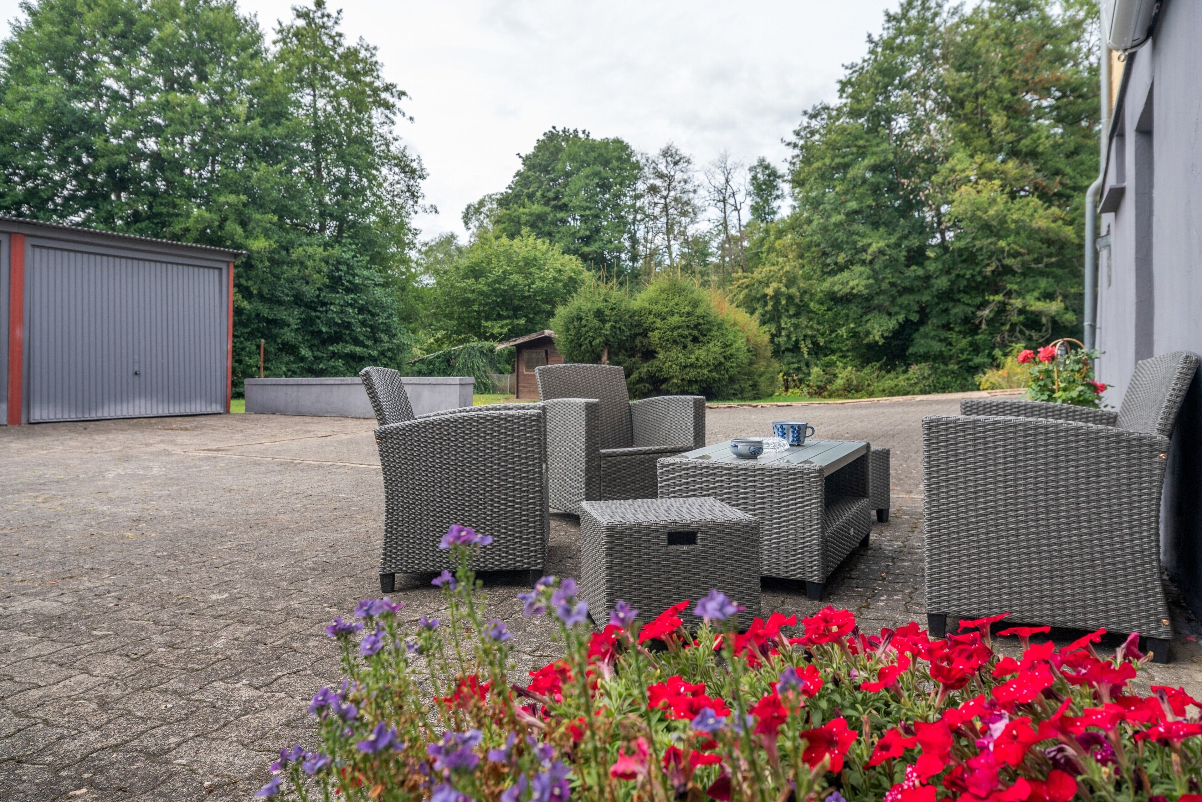 Un espace extérieur aménagé avec des meubles en osier et une petite table, entouré d’un jardin où poussent des fleurs rouges et violettes.