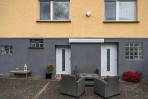 A residential building with a grey exterior, a white door, and a small patio area with wicker furniture and potted plants.