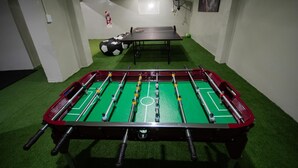 Una mesa de fútbol de mesa en una habitación con césped artificial verde, una mesa de ping pong y un balón de fútbol.