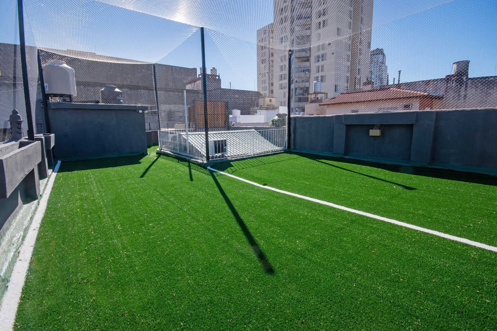 A rooftop with artificial green turf, a metal cage structure, and surrounding buildings.
