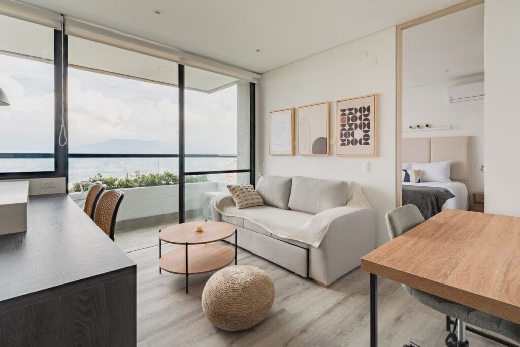 A modern living room with a sofa, a round coffee table, and a dining table. There are large windows offering a view of the city and mountains.