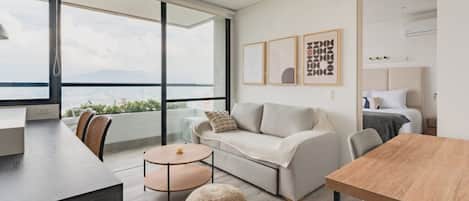 A modern living room with a sofa, a round coffee table, and a dining table. There are large windows offering a view of the city and mountains.