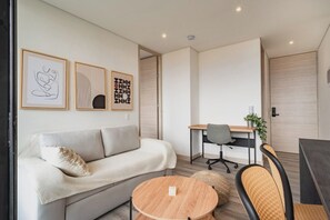 A modern living room with a sofa, a round wooden coffee table, and a desk with a chair. There are framed artworks on the wall and a plant on the desk. - ENZO Privé | Grand by Linares - Exclusive Corporate Apartment Suite in Medellín (Medellín)