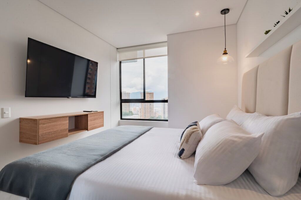 Een moderne slaapkamer met een groot bed, een wandgemonteerde televisie, een raam met uitzicht op de stad en een hanglamp.