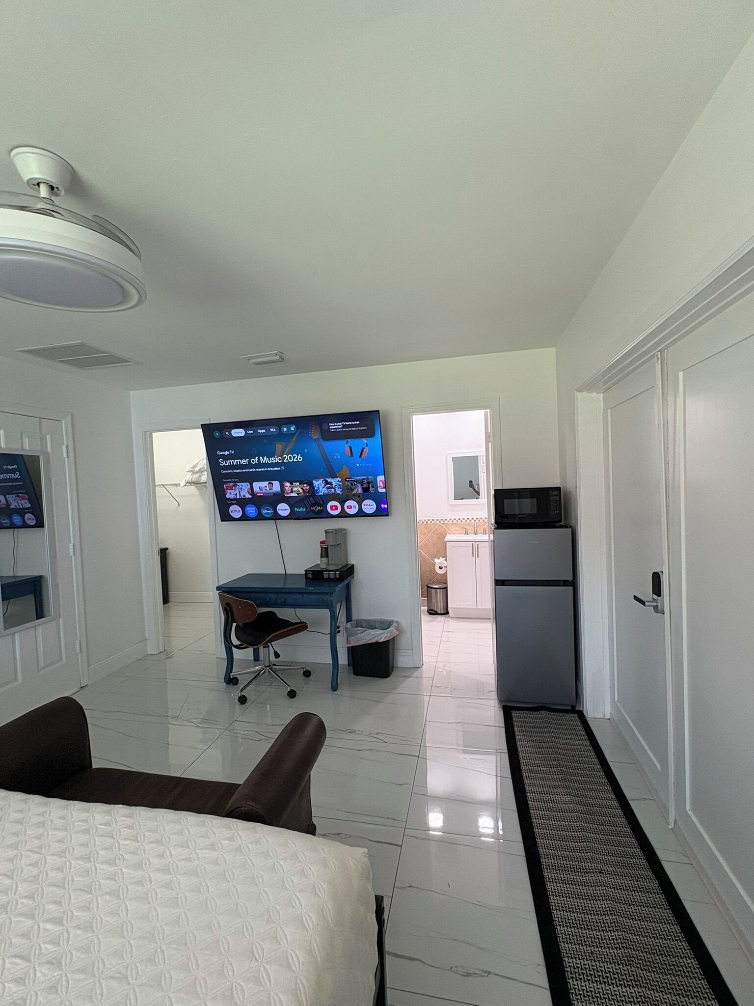 A hotel room with a bed, a desk with a computer, a TV mounted on the wall, and a kitchenette.