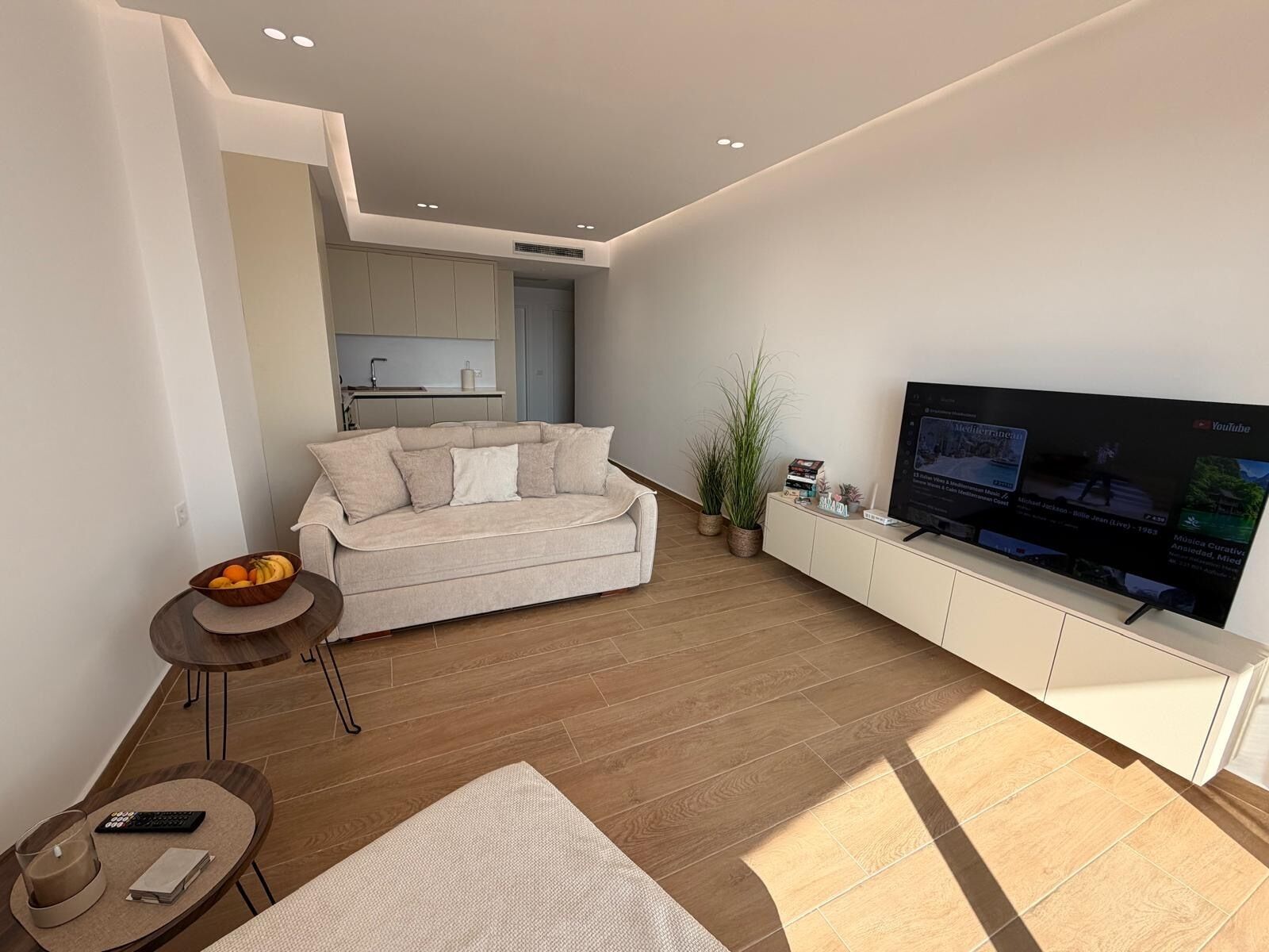 A modern living room with a sofa, a TV, a small table with a bowl of fruit, and a kitchen area in the background.