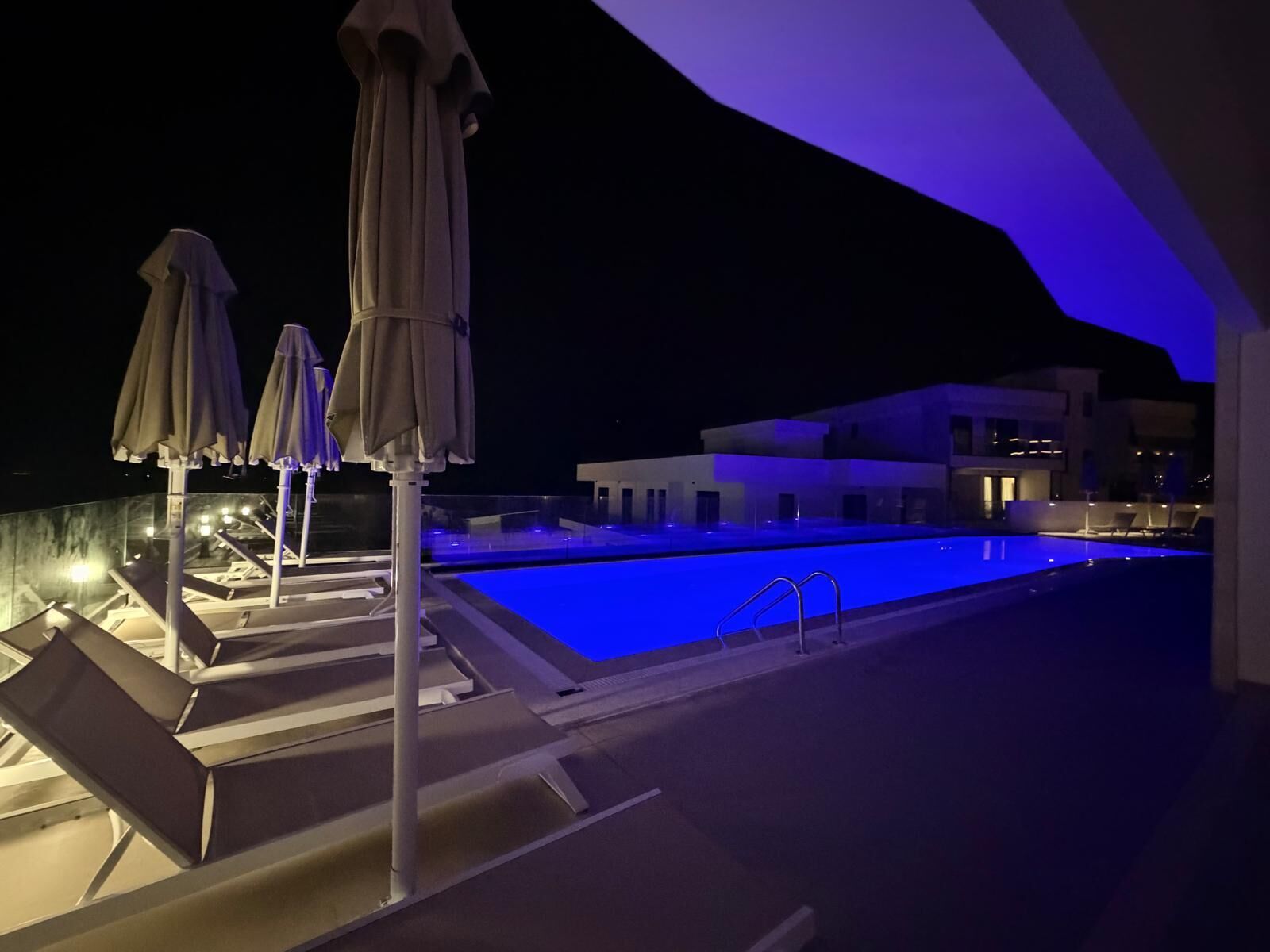 Area piscina notturna con poltrone a sdraio e ombrelloni, una piscina e un edificio moderno sullo sfondo.