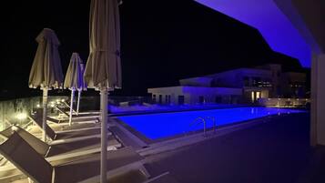 Area piscina notturna con poltrone a sdraio e ombrelloni, una piscina e un edificio moderno sullo sfondo.