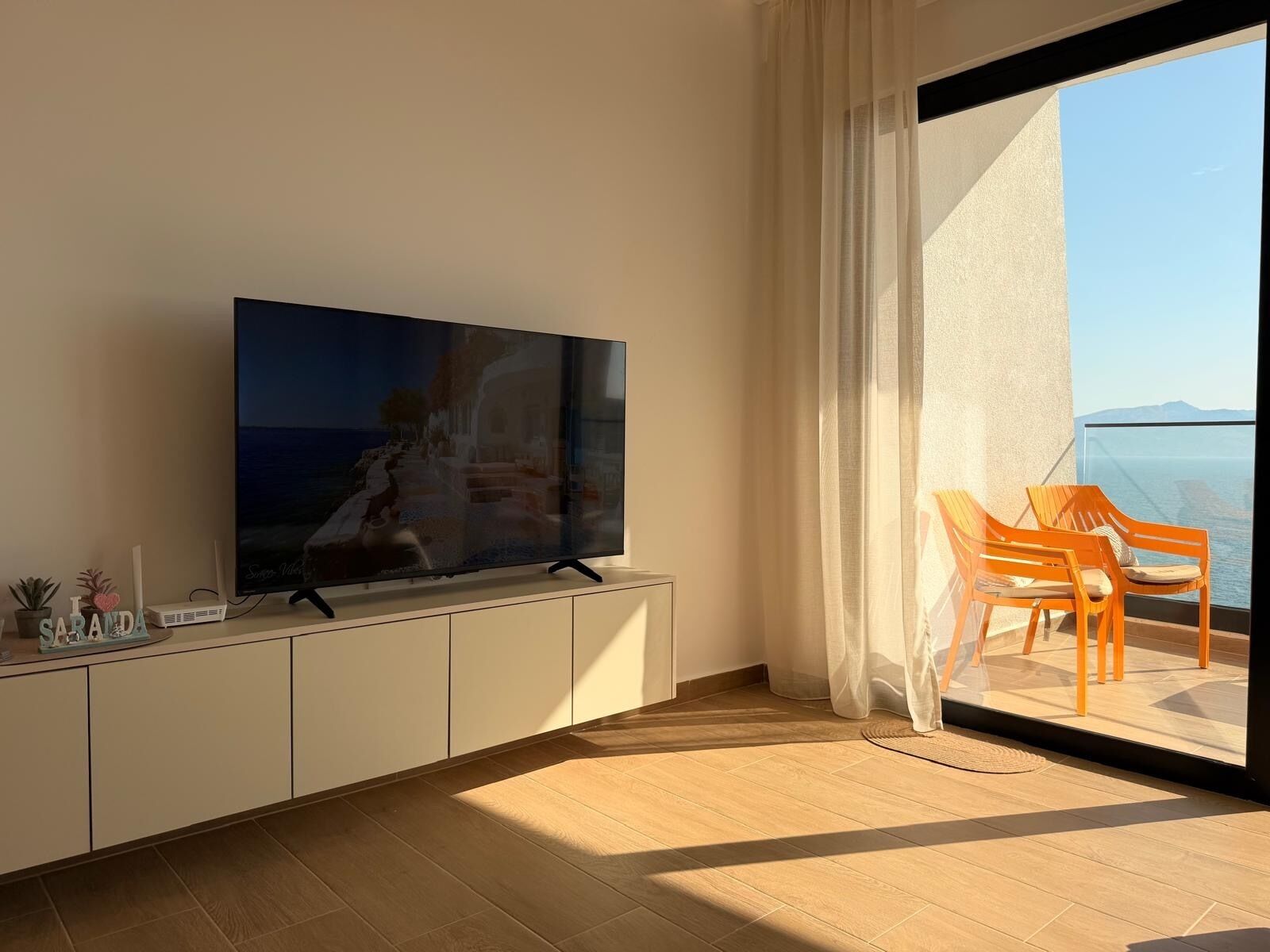 Un soggiorno moderno con TV a schermo piatto, pavimento in legno, un balcone con vista sul mare e un piccolo tavolo con oggetti decorativi.