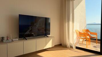 Un soggiorno moderno con TV a schermo piatto, pavimento in legno, un balcone con vista sul mare e un piccolo tavolo con oggetti decorativi.