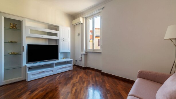 A living room with a flat-screen TV, built-in shelving, and a window with a view. - An elegant 75 sqm apartment, ideal for 4 people, located on the 6th and top floor of a building with an elevator (NOT accessible to people with disabilities).The Lodi metro station is just a 4-minute walk away, making it easy to get around the capital. (Rome)