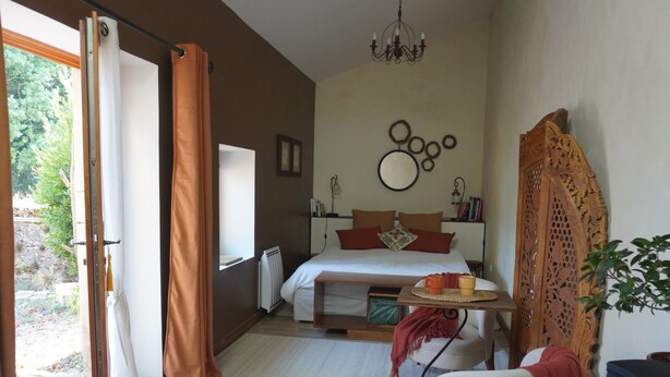 A bedroom with a bed, a bedside table, a chair, a window with curtains, a chandelier, and a view of greenery outside.