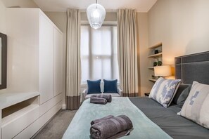 A hotel room with a bed, a bedside table, a lamp, a wardrobe, and a window with curtains. - Iconic 19th Century Marylebone Flat (London)