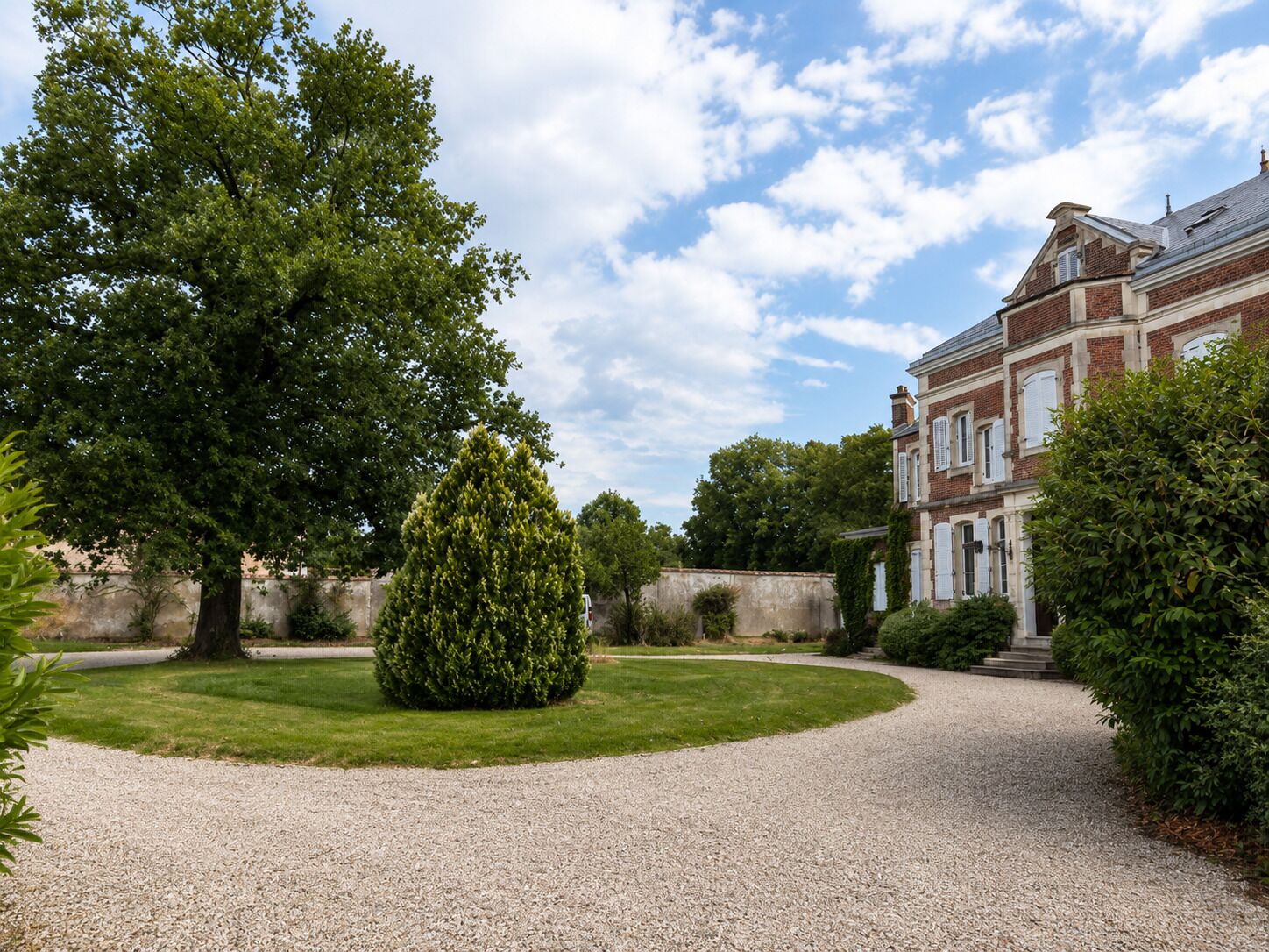 A gravel driveway leading to a building with a brick facade and a large tree in the foreground.