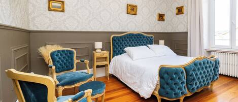 Een slaapkamer met een groot bed, twee fauteuils, een nachtkastje en drie ingelijste foto's aan de muur.